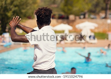 Black athletic hotel resort animator exercising in front of pool, view form behind blurred people background - water aerobic activity