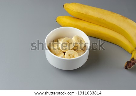 Two bananas and slices in a white plate on a gray background. Trending color of year 2021 Illuminating and Ultimate gray. Side view minimal still life.