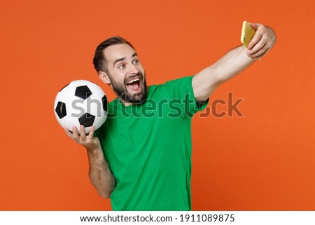 Excited young man football fan in green t-shirt cheer up support favorite team with soccer ball doing selfie shot on mobile phone isolated on orange background. People sport leisure lifestyle concept