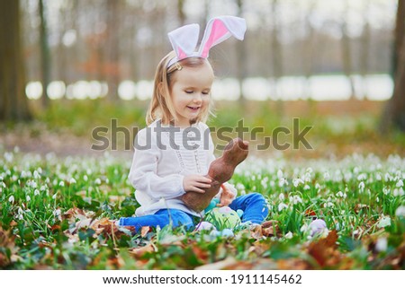 Girl wearing bunny ears playing egg hunt on Easter. Toddler sitting on the grass with many snowdrop flowers and eating chocolate bunny. Little kid celebrating Easter outdoors in park or forest