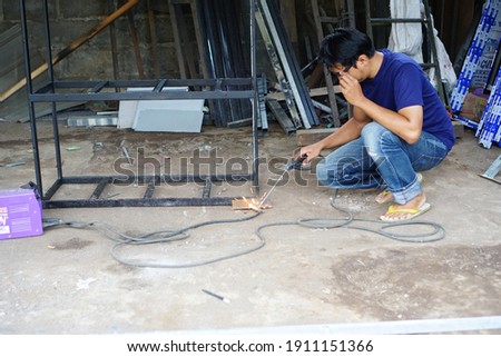 Squat male welder holding electric welder to weld iron frame against welding workshop background                      