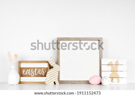 Mock up wood frame with Easter decor on a wood shelf. Shabby chic wood signs, eggs, bunnies. Square frame against a white wall. Copy space.