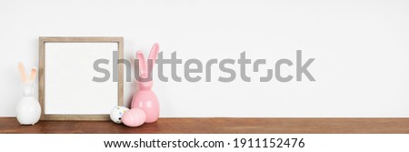 Mock up wooden frame with Easter Eggs and modern glass bunny decor on a wood shelf. Square frame against a white wall banner. Copy space.