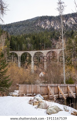 A photo of the famous Landwasser Viaduct Bridge in Switzerland with a house and a wooden bridge in the foreground. A cold day in late February just before the end of winter.