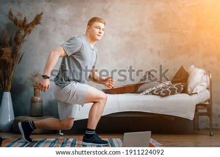 A young man goes in for sports at home, online workout from the laptop. The athlete lunges, watches a movie  in the bedroom, in the background there is a bed, a vase, a carpet.
