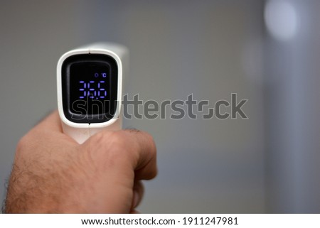 Non contact infrared forehead digital thermometer held by a young man with left hand with a defocused background. Closeup view Royalty-Free Stock Photo #1911247981