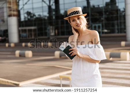 Cool young woman in boater and white dress smiles and looks into camera outside. Tourist holds passport and poses on crosswalk near airport. Royalty-Free Stock Photo #1911257254