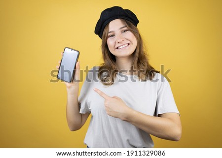 Beautiful young caucasian girl wearing french look with beret over isolated yellow background holding smartphone showing screen smiling happy pointing with hand and finger