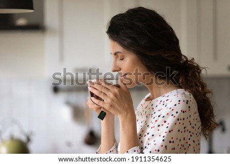 Happy millennial Caucasian woman hold mug drink enjoy hot black tea in the morning at home. Calm young female have warm beverage from cup, relax rest on weekend in cozy home. Hygge concept. Royalty-Free Stock Photo #1911354625