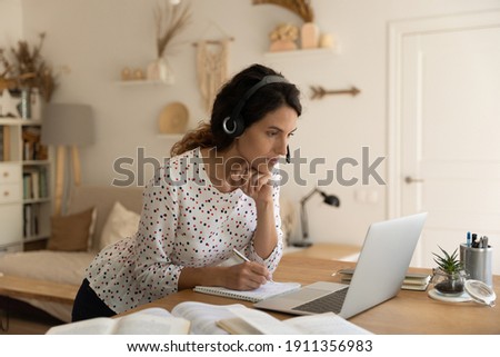 Focused millennial Caucasian female student in headphones look at laptop screen study distant make notes. Young woman work online on computer at home office, watch webinar or take training course. Royalty-Free Stock Photo #1911356983