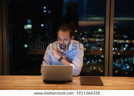 Young biracial male employee stay in office after working hours to study electronic documents on laptop screen. Thoughtful young guy corporate worker consider on difficult problem solution make choice Royalty-Free Stock Photo #1911366328