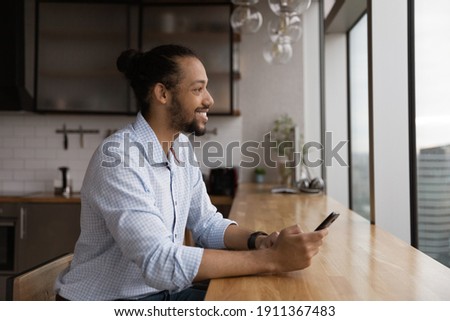 Smiling young african guy lean at desk hold mobile phone enjoy cityscape from picture window at modern kitchen. Positive black male renter owner of studio apartment dream distracted from cell screen