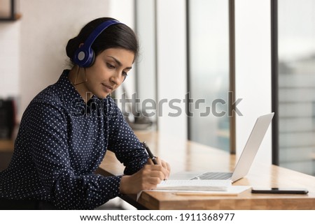 Focused millennial indian woman interpreter write translation from foreign language to english listen to audio record in headset. Attentive hindu female trainee hearing lecture course online take note