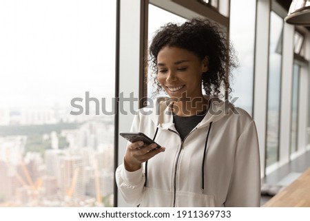 Smiling young african american lady spend free time at home in office use cellphone app. Content millennial black female stand by large picture window with splendid urban view messaging by cell online