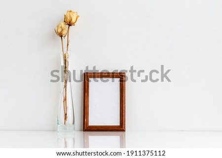A wooden frame with a white insert inside and white dried roses in a glass vase. Photo frame on a white wall background.
