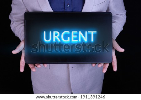 URGENT word, text written in neon letters on a laptop which is being held by a businessman in a gray suit.