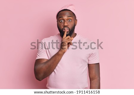Surprised dark skinned man with thick beard makes silence gesture keeps index finger over lips tells secret information dressed in casual t shirt and hat isolated over rosy background. Hush be silent