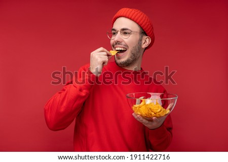Photo of happy man with beard in glasses and red clothing. Holds and eats a plate of chips, isolated over red background Royalty-Free Stock Photo #1911422716