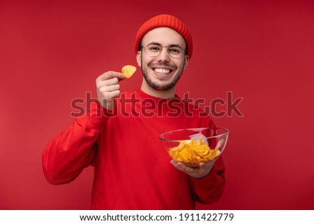 Photo of happy man with beard in glasses and red clothing. Holds and eats a plate of chips, isolated over red background Royalty-Free Stock Photo #1911422779