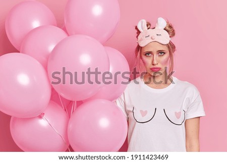 Photo of unhappy blonde woman with bright makeup has gloomy expression wears sleepmask and t shirt comes on pajama party holds bunch of balloons isolated over pink background. Spoiled holiday