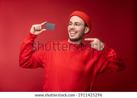 Photo of attractive man with beard in glasses and red clothing. Happy man holds and points at credit card, isolated over red background