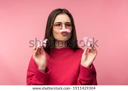 Photo of attractive lady in glasses plays with cotton candy, has fun. Wears casual pink sweater white pants isolated pink color background