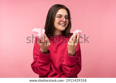 Photo of attractive lady in glasses plays with cotton candy, has fun. Wears casual pink sweater white pants isolated pink color background.
