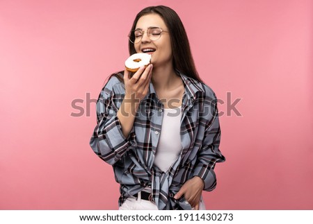 Photo of attractive lady holds eats donut sweet food, wear casual checkered shirt white undershirt isolated pink color background