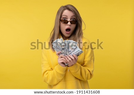 Photo of cute lady in glasses holds money in dollars, surprised and gape of salary. Wears casual yellow sweater, isolated yellow color background