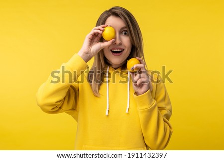 Photo of attractive lady poses in a cute way with lemons, cover one eye up with. Wears casual yellow hoody, isolated yellow color background