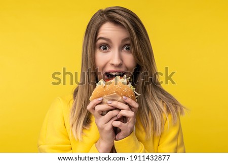 Photo of attractive lady poses in a happy way tries to bite burger. Wears casual yellow hoody, isolated yellow color background