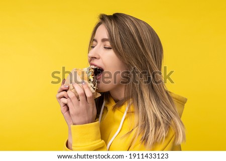 Photo of attractive lady poses in a surprised way bites burger. Wears casual yellow hoody, isolated yellow color background