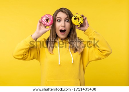Photo of attractive lady holds donuts in front of her eyes with opened mouth and shocked face. Wears casual yellow hoody, isolated yellow color background