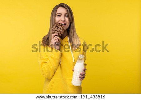 Photo of attractive lady bites cookie and holds milk, feels nice about dairy products. Wears casual yellow hoody, isolated yellow color background