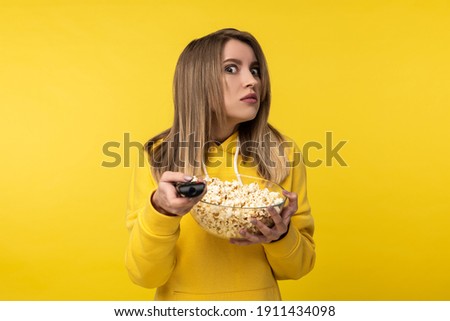 Photo of attractive lady holds tv remote and plate of popcorn, with disgruntled face tries to turn a channel. Wears casual yellow hoody, isolated yellow color background