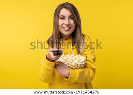 Photo of attractive lady holds tv remote and plate of popcorn, with happy face tries to turn a channel on. Wears casual yellow hoody, isolated yellow color background