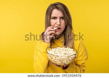 Photo of attractive lady holds plate of popcorn, eats it with disgruntled face. Wears casual yellow hoody, isolated yellow color background