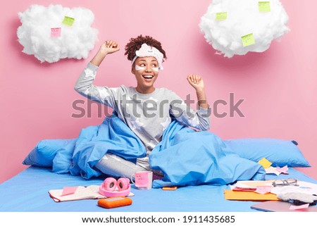 Happy dark skinned female student in pajama prepares university task at home stays in comfortable bed raises arms and dances celebrates good news undergoes beauty procedures poses at bedroom
