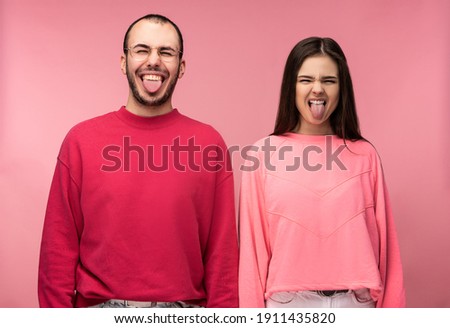 Photo of attractive man wih beard in red clothing and woman in pink grimace with tongue sticking out, isolated over pink background