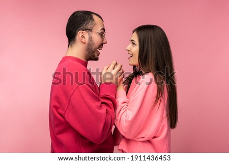 Photo of attractive man wih beard in red clothing and woman in pink holds for hands each other with mouth open. Couple looks happy, isolated over pink background