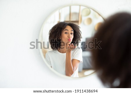 Great appearance, love yourself and perfect skin. Cheerful young african american lady makes air kiss and looks in reflection of round mirror in living room, on white wall, panorama, free space Royalty-Free Stock Photo #1911438694