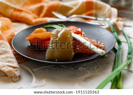 Pickled sweet bell pepper, cut into slices, multicolored vegetables in oil on a black matte plate and a stone white background. The dish is decorated with green leeks