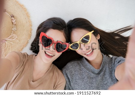 Portrait of two young Asian women wearing sunglasses in summer concept smiling and lying on the white floor to selfie take a photo.