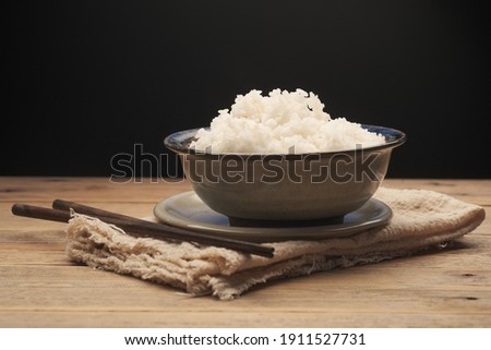 Cooked Thai jasmine rice in a ceramic bowl with chopsticks placed on an old wooden table against a black background. Royalty-Free Stock Photo #1911527731