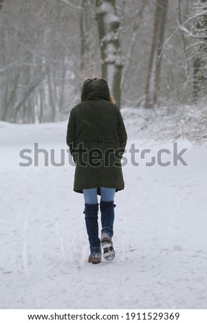 Blurred view on back of one unrecognizable woman walking alone on snow covered path in lonely german forest with bare trees during snowstorm in winter 