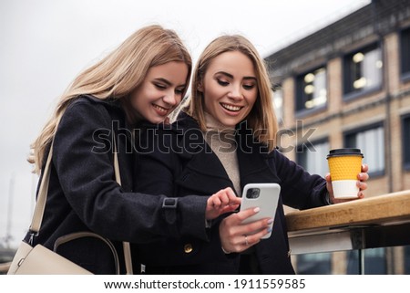 Two cute funny blonde girls makes selfie on their phone. They wear dark warm coats. Cold moody evening in european city