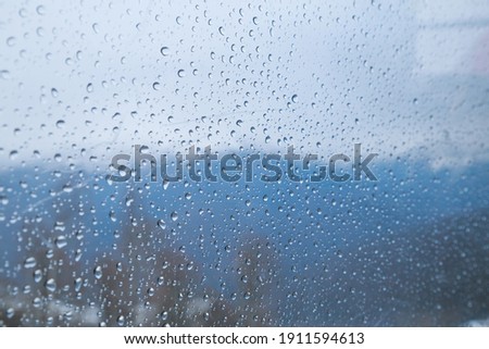 Rain drops on the scratched window glass background. Snowed mountains, trees, fog and blue cloudy sky behind.