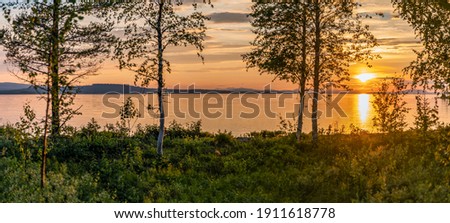 Scenic Panorama - Sunset over Umea river in mountains, summer sky with clouds highlighted by orange Sun. Blurry foreground with trees at coastline. Sunlight path on water. Storuman, Lapland, Sweden Royalty-Free Stock Photo #1911618778