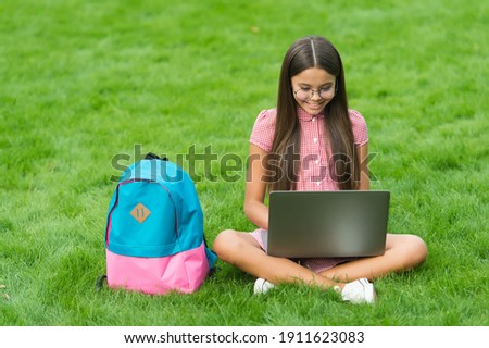smart happy kid wear glasses using laptop for doing school homework online sitting in park on green grass with backpack, education.