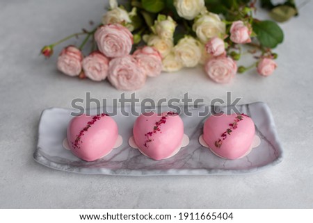 Three heart-shaped pink mirror glaze cakes decorated with gold leaf and dried roses with a rose bouquet on a stone background. Valentine's Day, Happy Birthday, Women's Day. Elegant greeting card
